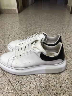 Alexander McQueen skor - Stilrena vita sneakers från Alexander McQueen med svart detalj på hälen och tjock sula. Skorna har klassisk snörning, perforerade sidor och är tillverkade i läder. Perfekta för dig som gillar en clean och modern look.