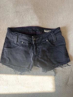 Svarta jeansshorts Lee - Snygga svarta jeansshorts från Lee med råa kanter och klassisk femficksdesign. Shortsen har bälteshällor, knapp- och dragkedjestängning samt coola slitdetaljer nertill. Perfekta för en avslappnad stil under varma dagar.