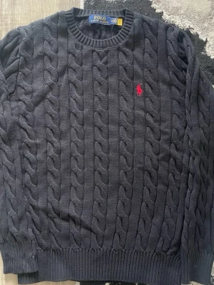  kabelstickad tröja Polo Ralph Lauren - Kabelstickad mörkblå tröja från Polo Ralph Lauren med rund halsringning och röd broderad logga på bröstet. Tröjan har ribbstickade muddar vid ärmslut och nederkant. Perfekt för en stilren och klassisk look.