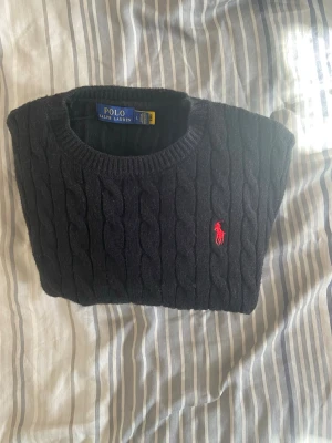 Svart kabelstickad tröja Polo Ralph Lauren - Kabelstickad svart tröja från Polo Ralph Lauren med rund halsringning och röd broderad logga på bröstet. Klassisk och tidlös design i mjukt material, perfekt för lager-på-lager-stil.