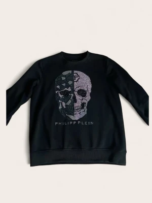 Svart sweatshirt från Philipp Plein - Cool svart sweatshirt från Philipp Plein med tryck och glittrande detaljer på bröstet. Tröjan har rund halsringning, långa ärmar och ribbade muddar. Perfekt för dig som gillar streetwear och vill sticka ut med en exklusiv look. Tröjan är i mycket bra skick.