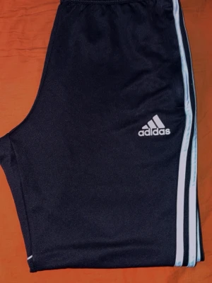 Svarta Adidas mjukisbyxor med ränder - Svarta mjukisbyxor från Adidas med klassiska vita ränder längs sidorna och Adidas-logga på låret. Byxorna har smal passform och resår i midjan. Tillverkade i ett mjukt och stretchigt material som passar perfekt för en avslappnad stil.