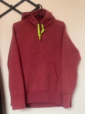 RevolutionRace röd hoodie - RevolutionRace röd hoodie med broderad logga på bröstet och en stor känguruficka framtill. Huvan har en cool neongul dragsko som sticker ut. Mjuk och skön bomullsblandning, perfekt för chill dagar.
