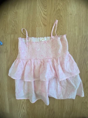 Ljusrosa volangig linnetopp - Supersöt ljusrosa linnetopp med smala axelband och volangdetaljer. Köpt från Gina barn storlek 152 men passar xs/xxs Toppen har smock upptill och ett skirt, lätt material med diskret mönster. Perfekt för varma dagar och ger en romantisk vibe.