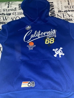 Blå hoodie California Glory - Blå hoodie med huva och känguruficka. Framsidan har tryck med texten 'California Glory 68', basketbollmotiv och olika patches. Ena ärmen har tryck med låga, 'Happy', 'smile' och spader-symbol. Perfekt för en avslappnad streetstyle.