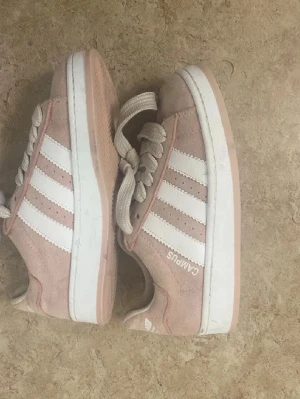 Adidas Campus rosa sneakers - Snygga Adidas Campus sneakers i ljusrosa mocka med klassiska vita ränder på sidorna och vit sula. Skorna har lågt skaft och grova vita snören. Perfekta för dig som gillar en clean och sportig stil.