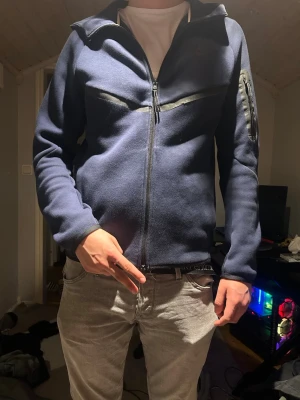 Mörkblå zip hoodie med ficka - Mörkblå Nike tech hoodie med dragkedja framtill och huva. Tröjan har en diskret ficka med dragkedja på ena ärmen och svarta detaljer längs bröstet. Strl S