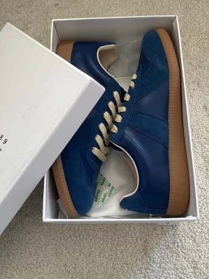 Mörk Blå Maison Margiela - Snygga blå sneakers från Maison Margiela med beige snörning och klassisk gummisula. Skorna har en mix av mocka och läderdetaljer som ger en stilren och modern look. Perfekta för dig som vill sticka ut med en ikonisk sneaker.