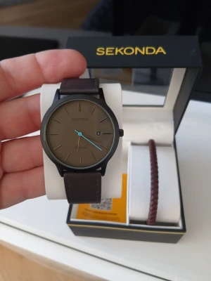 Sekonda  klocka med armband - Stilren klocka från Sekonda med brun läderrem och svart boett. Urtavlan är brun med minimalistiska markeringar och en blå sekundvisare. Kommer i originalask tillsammans med ett brunt flätat armband. Perfekt accessoar för en modern look.