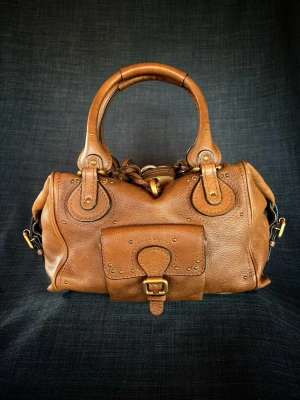 Chloé brun läder handväska - Snygg brun handväska från Chloé i äkta läder med rundade handtag, dekorativa nitar och en framficka med spänne. Väskan har guldfärgade metalldetaljer och en klassisk, lite boxig form. Perfekt för dig som gillar tidlös design och exklusiva material.