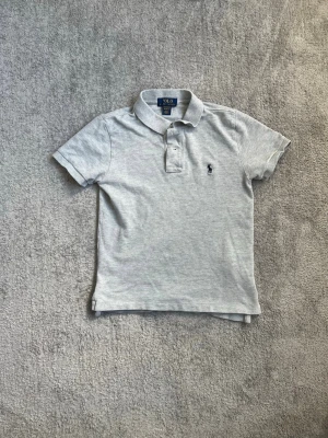 Ljusgrå pikétröja från Polo Ralph Lauren - Klassisk ljusgrå pikétröja från Polo Ralph Lauren med korta ärmar, krage och knappar framtill. Tröjan har den ikoniska broderade loggan på bröstet och är tillverkad i mjuk bomull. Perfekt för en clean och stilren look.