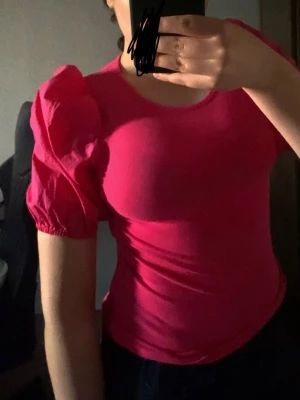 Rosa topp med puffärmar - Snygg rosa topp med trendiga puffärmar och rund halsringning. T-shirten är figurnära och har korta ärmar med resår vid ärmslut. Perfekt för dig som vill sticka ut med en färgstark look.