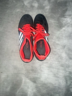 Adidas Predator fotbollsskor svart/röd - Snygga Adidas Predator fotbollsskor i svart med röda detaljer och vita ränder på sidorna. Skorna har röda snören, stickad strumpliknande öppning och dobbar för bra grepp på planen. Perfekta för dig som vill ha kontroll och stil på fotbollsplanen.
