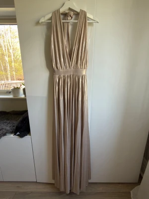 Champagnefärgad långklänning Goddiva - Elegant långklänning från Goddiva i skimrande champagnefärg. Klänningen har djup v-ringning, öppen rygg och bred midjeresår och ett mjukt, glansigt material som faller snyggt. 