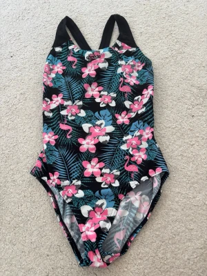 Blommig baddräkt från Speedo - Färgglad baddräkt från Speedo med tropiskt blommönster i rosa, blått och vitt samt flamingos. Svarta axelband och öppen rygg ger en sportig look. Perfekt för strand eller pool!
