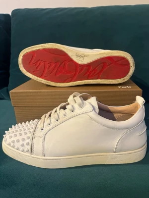  Christian Louboutin skor - 🛑 Vita sneakers från Christian Louboutin i läder med ikoniska röda sulor och nitar på tån. 🛑Storlek 45 i ganska bra skick, finns inga större defekter.🛑Hör av dig vid funderingar!!!