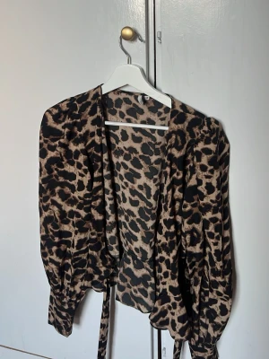 Leopardmönstrad omlottblus - Snygg omlottblus med leopardmönster i beige och svart. Blusen har långa ärmar med lätt puff och knytband i midjan. Tillverkad i ett mjukt, glansigt tyg som ger en lyxig känsla. Perfekt för att lyfta vilken outfit som helst.