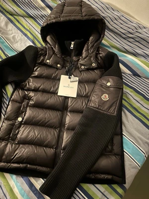 Svart dunväst med huva från Moncler - Snygg svart cardigan från Moncler med stickade svarta ärmar och huva. Jackan har dragkedja, tryckknappar och en ficka med Moncler-logga på ärmen. Perfekt för lager-på-lager och streetstyle.