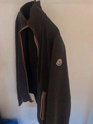 Svart Moncler ziptröja med ränder - Snygg svart ziptröja från Moncler med klassiska ränder i rött, vitt och blått längs dragkedjan och vid fickorna. Tröjan har hög krage och Moncler-logga på ärmen. Perfekt för en stilren och sportig look. Söndrig zip går ej att knäppa. Pris kan diskuteras. Äkta! 