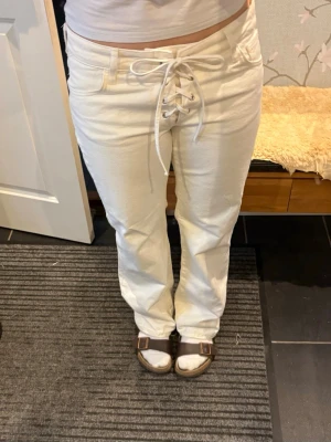 Vita jeans med snörning i midjan - Säljer ett par vita jeans med snygg snörning framtill i midjan. Byxorna har raka ben, klassiska fickor och coola detaljer som ger en unik look. Jag själv bär i vanliga fall st 38 och jeansen passar mig perfekt. 