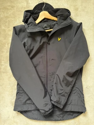 Svart vindjacka från Lyle & Scott - Snygg svart vindjacka från Lyle & Scott med gul logga på bröstet. Jackan har huva, dragkedja framtill och elastiska muddar vid ärmsluten. Tillverkad i lätt och slitstarkt material, perfekt för blåsiga dagar. Inga defekter alls eller problem 
