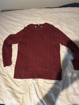 Vinröd kabelstickad tröja Tommy Hilfiger - Snygg vinröd kabelstickad tröja från Tommy Hilfiger med rund halsringning och diskret logga på bröstet. Tröjan har ribbade muddar och nederkant, samt klassiska stickade detaljer. Perfekt för dig som gillar stilrena och tidlösa plagg.