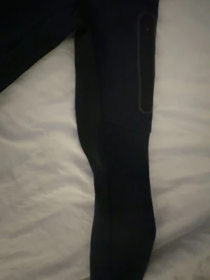 Svart Nike Tech med huva - Svart sportig träningsjacka med hel dragkedja och huva. Jackan har diskreta reflexdetaljer på bröstet och en ficka med dragkedja på ärmen. Perfekt för löpning eller utomhusträning. Snygg och funktionell design.