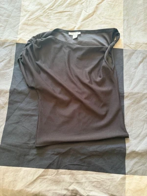 Svart topp från Amisu med drapering - Snygg svart topp från Amisu med lätt draperad axel och enkel, stilren design. Toppen är ärmlös och har en mjuk känsla, perfekt att matcha med jeans eller kjol. Tillverkad i ett stretchigt material som sitter skönt på kroppen.