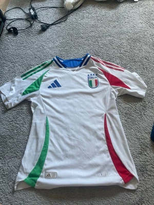 Italien Adidas fotbollströja vit - Säljer en officiell Italien fotbollströja från Adidas i vitt med gröna och röda detaljer på ärmar och sidor. Tröjan har Italiens landslagsmärke på bröstet och blå krage. Tillverkad i lätt och ventilerande material, perfekt för träning eller match. 