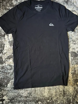 Svart Quiksilver t-shirt i bomull - Svart t-shirt från Quiksilver med liten logga på bröstet. Klassisk rund halsringning och korta ärmar. Tillverkad i mjuk bomull, perfekt för en avslappnad stil.