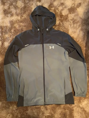 Grå-Under Armour vindjacka - Snygg vindjacka från Under Armour i grått med hel dragkedja och huva. Jackan har logga på bröstet, justerbar huva och diskreta reflexdetaljer. Perfekt för blåsiga dagar och sportiga outfits. Priset går och diskutera vid snabbaffär