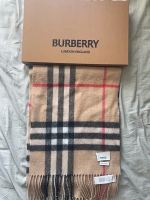 Beige rutig halsduk från Burberry - Klassisk beige halsduk från Burberry i mjuk ull med det ikoniska rutmönstret i svart, vitt och rött. Halsduken har fransar i kanterna och kommer med originalförpackning och certifikat. Perfekt accessoar för att lyfta din outfit. Taggar, box, precis allting ingår.