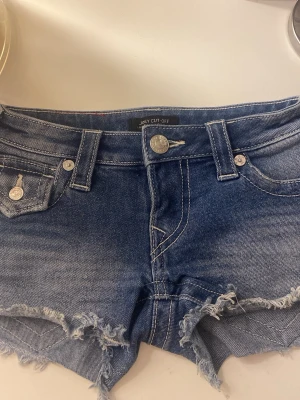 True Religion blå jeansshorts - Blå jeansshorts från True Religion med låg midja, fransig och rå kant nertill samt klassiska fickor fram och bak. Shorts i bomull med slitna detaljer och silverfärgade knappar. Perfekt för en avslappnad och trendig sommarstil! Köpta för 1000kr säljer för 800 kontakta om ändring av pris kan tänka mig o sänka. Bara användt 3 gånger ungefär, skriv om frågor!👍💕