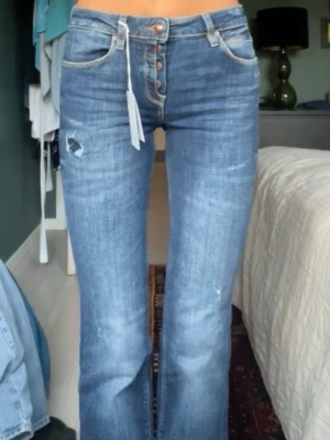 Blå bootcut jeans från Guess - Säljer ett par mörkblå bootcut jeans från Guess med slitna detaljer. Jeansen har knäppning fram, med fina knapp detaljer. Materialet är stretchigt denim som sitter snyggt och bekvämt. De är långa och passar mig som är 178.                  Storlek: 25, Innerbenslängd: 88 cm