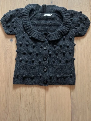Mörkgrå stickad kofta med puffärm - Kortärmad mörkgrå stickad kofta med stora knappar framtill och dekorativa bollar över hela plagget. Koftan har två fickor framtill, ribbad krage och en mysig, chunky känsla. Perfekt för lager på lager och en cool, trendig look.