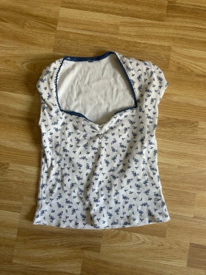 Vit blommig ribbad topp - Söt vit ribbad topp med blått blommönster och mörkblå kant vid halsen. Toppen har korta ärmar och en lätt v-ringad urringning. Perfekt för dig som gillar romantisk och feminin stil.