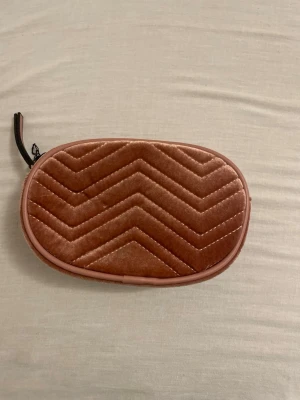 Rosa quiltad handväska i sammet - Söt liten handväska i rosa sammet med quiltat sicksackmönster. Väskan har oval form och dragkedja upptill med svart dragkedjesnodd. Perfekt för att bära småsaker och accessoarer. Enkel men trendig design som sticker ut. Axelremmen ligger innuti väskan då den är helt ny och oanvänd. 