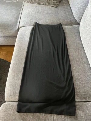 Svart långkjol i mjukt material - En enkel och stilren svart långkjol i mjukt, stretchigt material. Kjolen har rak passform och är perfekt för dig som gillar minimalistisk stil. Inga synliga detaljer eller mönster, bara clean och klassisk look. Lappen är bortklipps så inte säker var det är ifrån, om jag får gissa, H&M