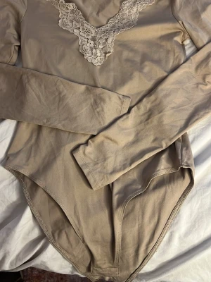Beige body med spetsdetalj H&M - Säljer en beige långärmad body från H&M med v-ringning och snygg spetsdetalj framtill. Tillverkad i mjukt stretchigt material som sitter skönt på kroppen. Perfekt att styla med jeans eller kjol för en trendig look.