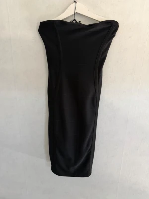 Svart axelbandslös långklänning - Elegant svart långklänning med axelbandslös design och figurnära passform. Klänningen har en stilren och minimalistisk look, perfekt för dig som gillar klassiska plagg. Tillverkad i ett  tight material som formar sig efter kroppen.