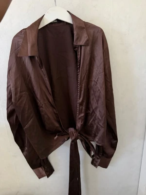 Brun knytblus från Bikbok - Snygg brun blus i glansigt material från New Look. Blusen har långa ärmar med manschetter, klassisk krage och knytning framtill för en trendig look. Perfekt att styla med höga jeans eller kjol.