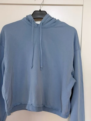 Ljusblå hoodie med vågig kant - En hoodie med croppad passform och råa, rullade kanter nertill, och vid ärmsluten. Justerbar huva med dragsko, sänkta axlar och en kortare längd.  Använd sparsamt! Inga skavanker! Säljer en grå också.  Nypris 349 kr 