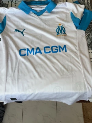 Marseille matchtröja Puma vit/blå - Säljer en vit Marseille matchtröja från Puma med blå detaljer och klubbmärke på bröstet. Tröjan har sponsortryck 'CMA CGM' och klassisk V-ringning. Perfekt för dig som älskar fotboll och vill stötta ditt lag med stil.