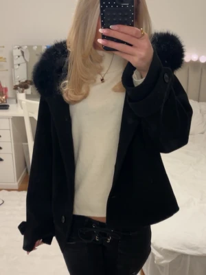 Svart jacka me päls - Svart kappa/jacka från Gina i Stl M men passar mig som brukar ha XS-S. Använd men inga defekter. Den har stora fickor i sidan, knappar framtill och en luva med päls som även är avtagbar. Passar både till vintern men även en vår eller höst jacka❤️ 