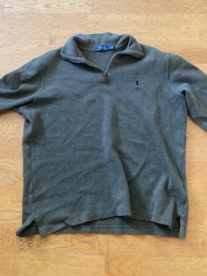 Olivgrön tröja från Polo Ralph Lauren - Olivgrön långärmad tröja från Polo Ralph Lauren med halv dragkedja och liten blå logga broderad på bröstet. Tröjan har hög krage och är tillverkad i mjukt material, perfekt för lager på lager. Klassisk och stilren design. Tröjan är i storlek m. Den är använd men det finns inga tydliga defekter på den. 
