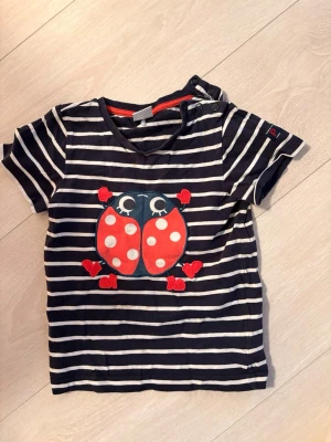 Randig t-shirt med nyckelpiga Polarn O. Pyret - Mörkblå och vit randig t-shirt från Polarn O. Pyret med korta ärmar och en söt applikation av en nyckelpiga framtill. T-shirten har tryckknappar vid axeln för enkel påklädning och är gjord i mjuk bomull.