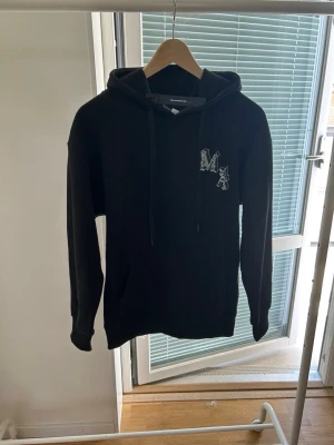 Svart hoodie från Amiri med tryck - Svart hoodie från Amiri med coolt vitt tryck på ryggen som föreställer en ängel och texten 'AMIRI'. Passar som XS. På bröstet finns ett mindre vitt 'MA'-tryck. Hoodien har huva med dragsko och är tillverkad i mjukt material. Perfekt för en streetwear-look.