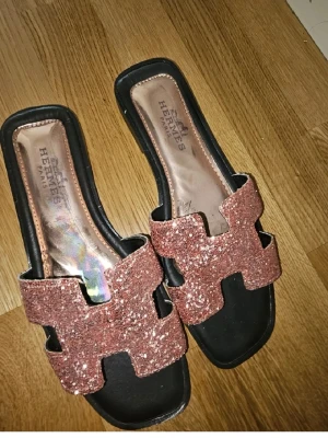 Glittriga sandaler  - Säljer ett par snygga sandaler med glittriga, roséguldiga remmar i klassisk H-design. Sandalerna har svart sula och öppen tå, perfekt för dig som vill sticka ut med lite extra bling. Passar till många olika outfits och ger en lyxig känsla.