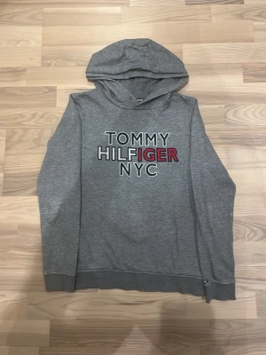 Grå hoodie Tommy Hilfiger NYC - Snygg grå hoodie från Tommy Hilfiger med trycket 'TOMMY HILFIGER NYC' på bröstet i vitt, svart och rött. Klassisk huva och ribbade muddar. Perfekt för en chill och stilren look. Materialet känns mjukt och bekvämt.