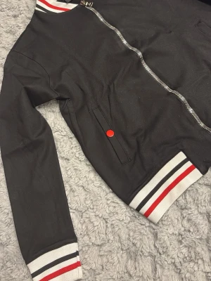 Svart jacka från Moncler - Svart jacka från Moncler med dragkedja framtill, vita och röda ränder på muddar och krage samt en röd knapp på fickan. Jackan har klassisk bomberstil och är tillverkad i ett slätt tyg. Perfekt för en sportig och trendig look.